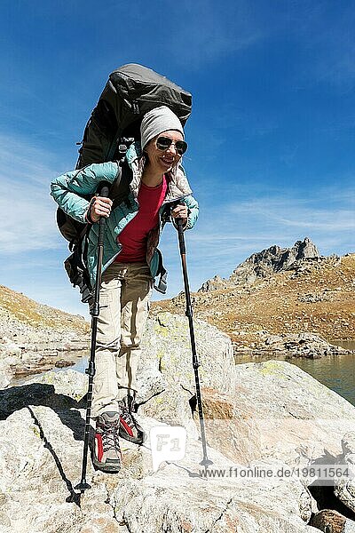 Schlankes und sympathisches Wandermädchen mit Sonnenbrille und blaür dünner Daunenjacke mit Rucksack und Trekkingstöcken steigt auf einen hohen Felsen vor dem Hintergrund von Felsen und einem hohen Bergsee