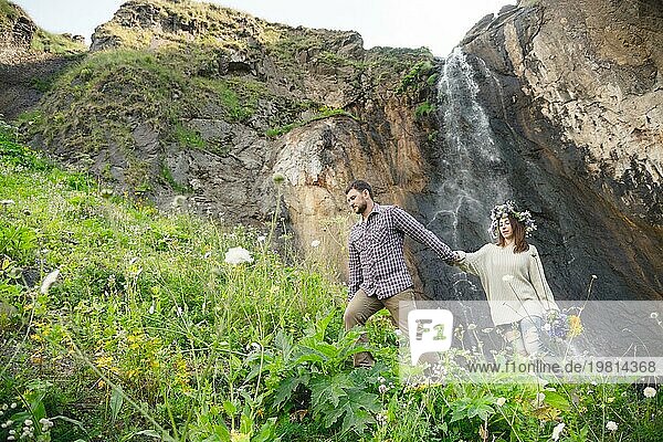 Hipster mit einem Bart führt seine Braut in der Natur vor dem Hintergrund eines Wasserfalls  der von einem Felsen fällt