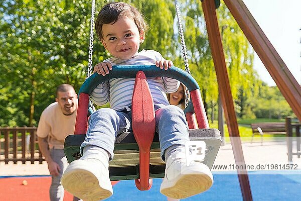 Frontalansicht von Eltern  die ein kleines Kind auf dem Spielplatz schaukeln  an einem sonnigen Tag