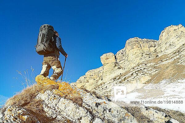 Ein Rucksacktourist mit einem großen Rucksack und Stöcken steigt in der Wintersaison vor dem Hintergrund epischer Felsen zum Felsen auf. Das Konzept des Bergtourismus das ganze Jahr über