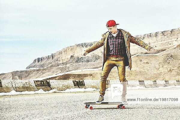 Stylish happy Junger Mann in Mütze und Hose Jogger rollen auf einem Longboard eine Bergstraße hinunter  genießen das Leben