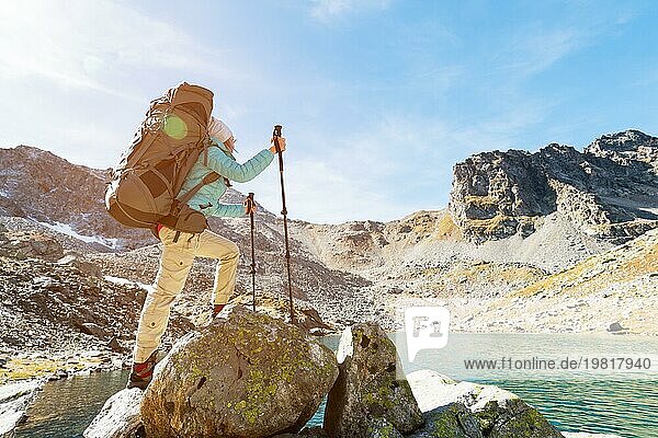 Schlankes und sympathisches Wandermädchen mit Sonnenbrille und blaür dünner Daunenjacke mit Rucksack und Trekkingstöcken steigt auf einen hohen Felsen vor dem Hintergrund von Felsen und einem hohen Bergsee