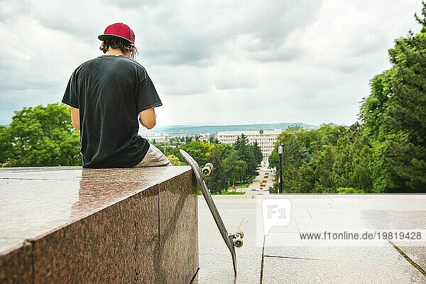 Der langhaarige Skater mit Mütze und TShirt sitzt mit dem Rücken zur Kamera und denkt neben dem Skateboard vor dem Hintergrund der Landschaft