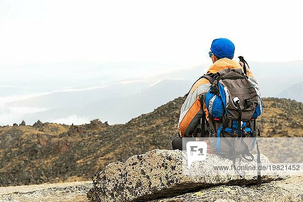Ein einsamer Tourist mit einem Rucksack und Stöcke für die Bewegung in den Bergen. In Sonnenbrille und mit einem Rucksack Rest sidiya auf einem Stein. Blick von hinten