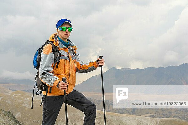 Ein einsamer Tourist mit Rucksack und Sonnenbrille genießt die Aussicht hoch in den Bergen des Kaukasus  wo es kein Gras gibt  das Dorf und den Schnee bei Sonnenuntergang
