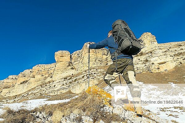 Ein Rucksacktourist mit einem großen Rucksack und Stöcken steigt in der Wintersaison vor dem Hintergrund epischer Felsen zum Felsen auf. Das Konzept des Bergtourismus das ganze Jahr über
