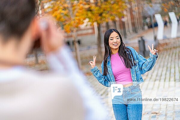 Chinesische junge Frau gestikuliert den Sieg  während ein Freund auf der Straße ein Foto mit einer Kamera macht