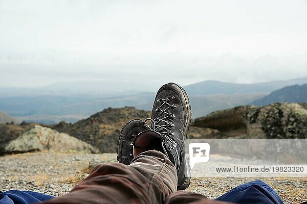 Beine eines Mannes  der sich in Stiefeln für eine Bergwanderung ausruht  vor dem Hintergrund von Bergen und Tälern mit lärmenden Wolken Wanderstiefel in den Bergen. Zeit zum Ausruhen. Freiheit Konzept