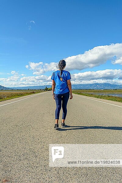Ein Reisender in legerer Kleidung  der eine gepflasterte Straße in Richtung des Horizonts in einer ruhigen Umgebung entlanggeht  Rückenansicht einer blau gekleideten Latina  die die Straße im Naturpark Ebrodelta entlanggeht  Tarragona  Katalonien  Spanien  Europa