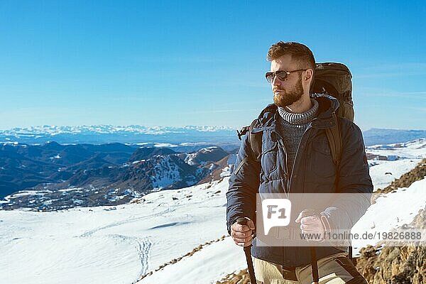 Ein Hipster Reisender mit Bart und Sonnenbrille in der Natur. Ein Mann wandert in den Bergen mit einem Rucksack und skandinavischen Wanderstöcken vor dem Hintergrund einer Berglandschaft und blauem Himmel. Reisen Lifestyle Abenteuer Outdoor Erholung Outdoor Sport