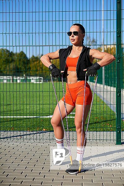 Schönes junges Fitnessmädchen wärmt sich vor dem Training im Stadion mit einem Springseil auf. Attraktive schlanke Brünette im orangefarbenen Trainingsanzug. Aktiver Lebensstil