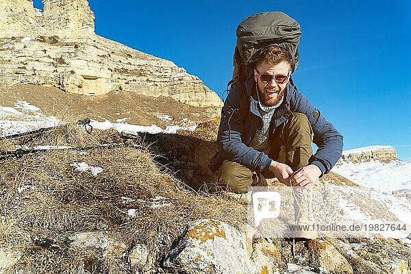 Porträt eines glücklich lachenden Hipster Reisenden mit Bart und Sonnenbrille sitzt und bindet Schnürsenkel in der Natur. Ein Mann wandert in den Bergen mit einem Rucksack und skandinavischen Wanderstöcken im Hintergrund einer Berglandschaft. Reisen Leben