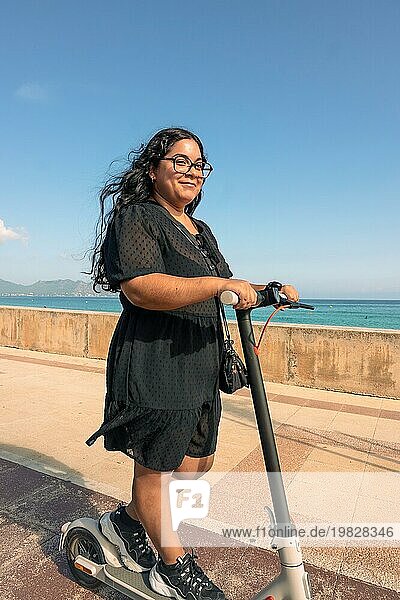 Eine fröhliche Frau fährt an einem sonnigen Tag mit einem Elektroroller am Meer entlang