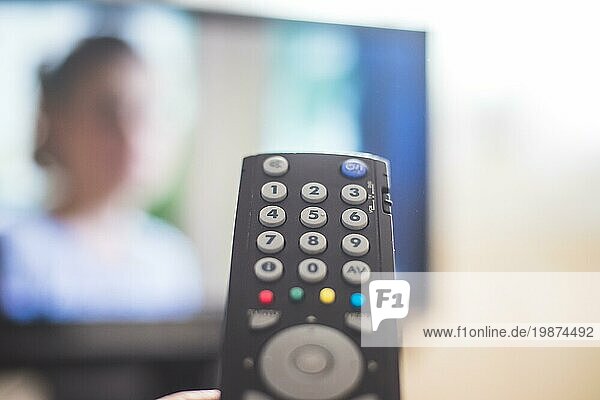 Eine TV Fernbedienung in der Hand  im Vordergrund  der Fernseher im verschwommenen Hintergrund. Streaming
