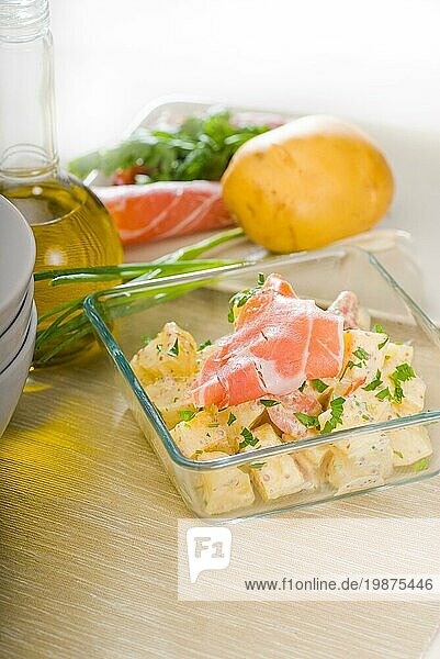 Frischer hausgemachter Parmaschinken und Kartoffelsalat  mit rohen Zutaten auf einem Tisch mit Schüsseln und Geschirr  Foodfotografie