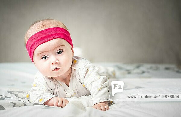 Adorable 6 Monate alt Baby Mädchen Kleinkind auf einem Bett auf dem Bauch mit dem Kopf nach oben in die Kamera mit ihren großen Augen. Natürliches Schlafzimmer Licht