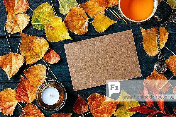 Eine leere braune Handwerk Grußkarte oder Einladung mit einer Tasse Tee  Herbstblätter und eine Kerze  ein Herbst Briefpapier Mockup  Overhead Flachlegung Schuss auf einem dunkelblauen hölzernen Hintergrund