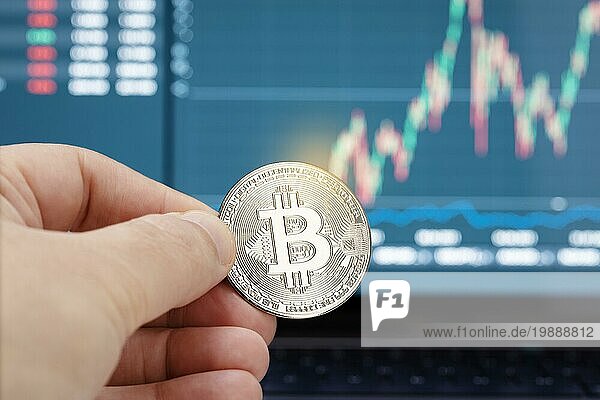 Hand hält eine Bitcoinmünze und Chart auf Laptop Display im Hintergrund. Cryptocurrency Handel oder Investitionen Konzept