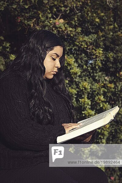 Junge Frau  sitzend und aufmerksam ein Buch lesend im Garten vertikale Halbkörper Seitenansicht Foto