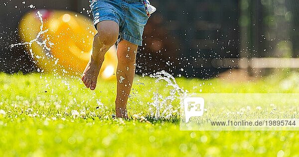 Fröhliches Kind läuft durch naßen Rasen und spritzt mit Wasser. Kindheit Konzept