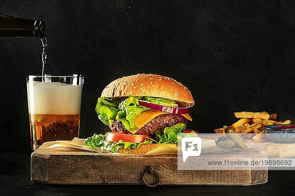 Burger und Bier. Hamburger mit Rindfleisch  Käse  Zwiebeln  Tomaten und grünem Salat  eine Seitenansicht auf einem dunklen Hintergrund mit Kopierraum  Gurken und Pommes frites. Selektiver Fokus