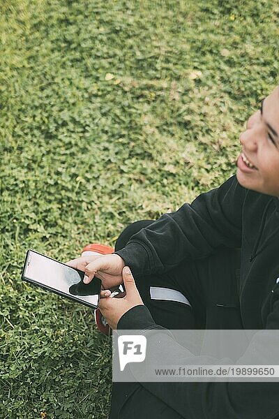 Ein fröhlicher Teenager lateinamerikanischer Abstammung lächelt  während er auf seinen Smartphonebildschirm schaut. Die entspannte Körperhaltung und der fröhliche Ausdruck des Jungen vermitteln ein Gefühl der Zufriedenheit. Der Hintergrund ist unscharf  um die Aufmerksamkeit auf den Jungen und sein Telefon zu lenken