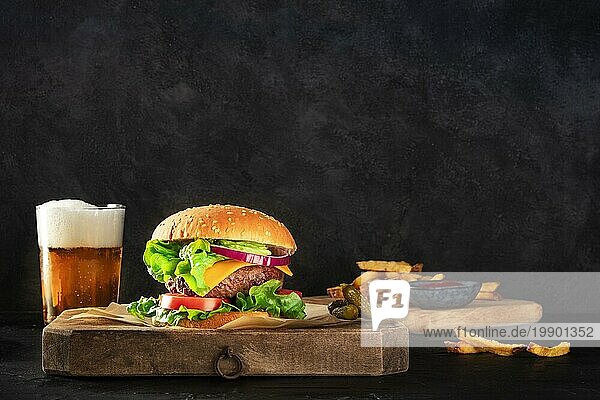Burger und Bier. Hamburger mit Rindfleisch  Käse  Zwiebel  Tomate und grünem Salat  eine Seitenansicht auf einem dunklen Hintergrund mit Gurken und Pommes frites  mit einem Platz für Text. Selektiver Fokus