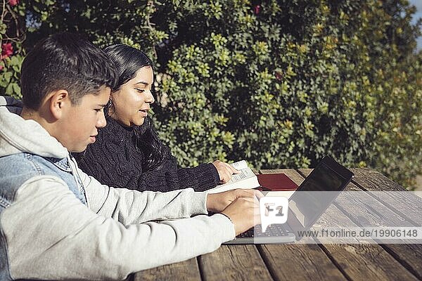 Zwei Kinder lernen mit einem Laptop und Büchern auf einem Holztisch im Freien an einem sonnigen Tag