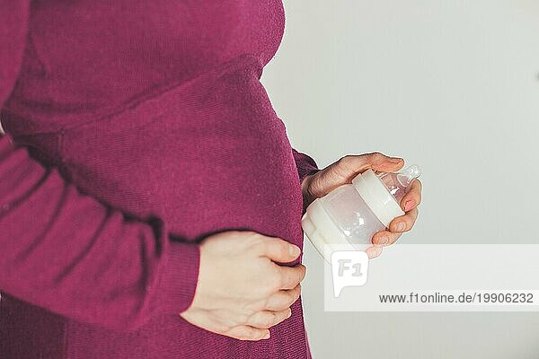 Nahaufnahme einer jungen Mutter  die eine Babyflasche auf ihrem schwangeren Bauch hält  Kaukasierin  magentafarbenes Kleid