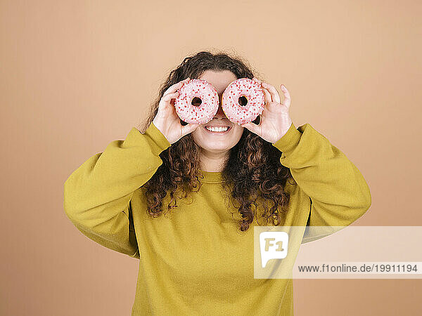Playful woman holding doughnuts over eyes against peach background