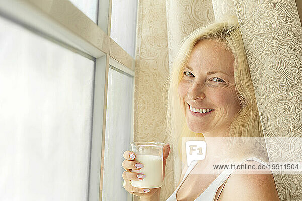 Smiling woman holding glass of milk near window at home