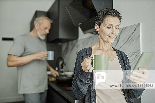 Woman using smartphone while her husband is cooking lunch in background