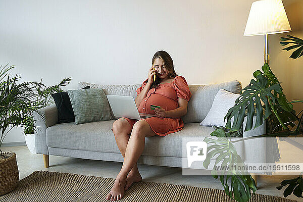 Smiling pregnant woman talking on smart phone sitting with laptop at home