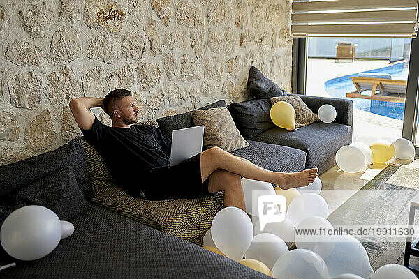Freelancer with laptop sitting amidst balloons on sofa at villa