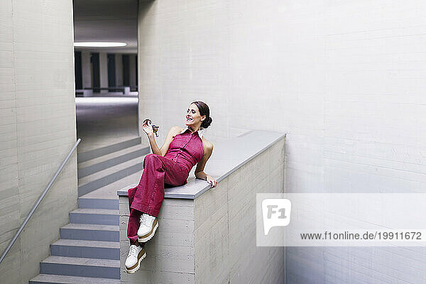 Smiling woman wearing pink jumpsuit and sitting on wall near staircase