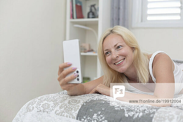 Happy woman doing video call through smart phone lying on bed at home