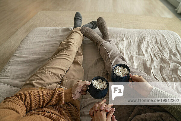 Couple holding mugs of hot chocolate with marshmallows on bed at home
