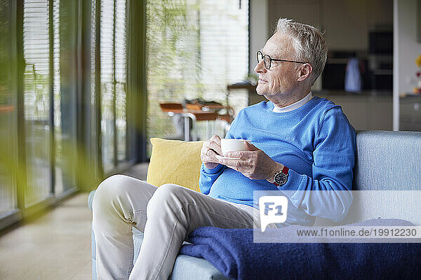 Thoughtful senior man sitting on couch at home with cup of coffee