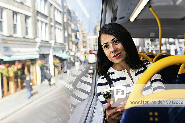 Smiling woman holding smart phone and looking through window in bus