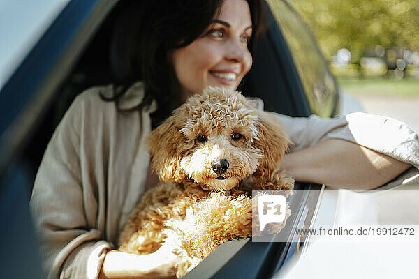 Smiling woman holding poodle dog and leaning out of window in car