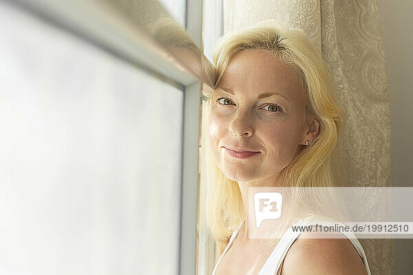 Smiling blond woman leaning on window at home