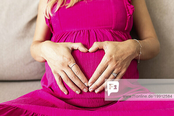 Pregnant woman wearing pink dress and gesturing heart shape on stomach