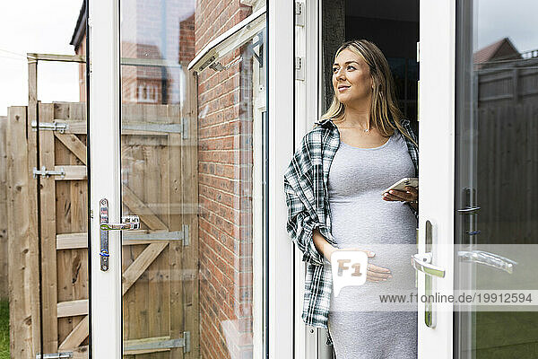 Smiling pregnant woman standing with hand on stomach and holding smart phone at balcony door