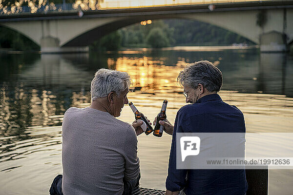 Senior friends toasting beer bottles near water at sunset