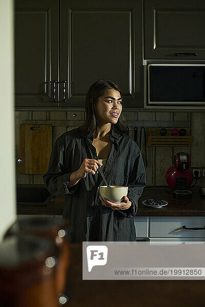 Smiling woman holding bowl of breakfast and standing in kitchen at home