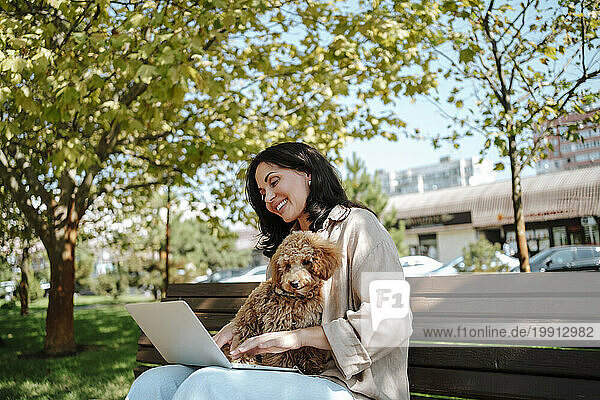 Smiling freelancer using laptop with poodle dog sitting on lap at park