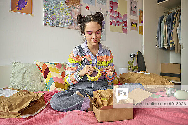 Young woman with adhesive tape packing box on bed at home