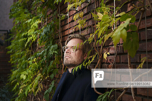 Contemplative mature businessman leaning on brick wall overgrown with plants
