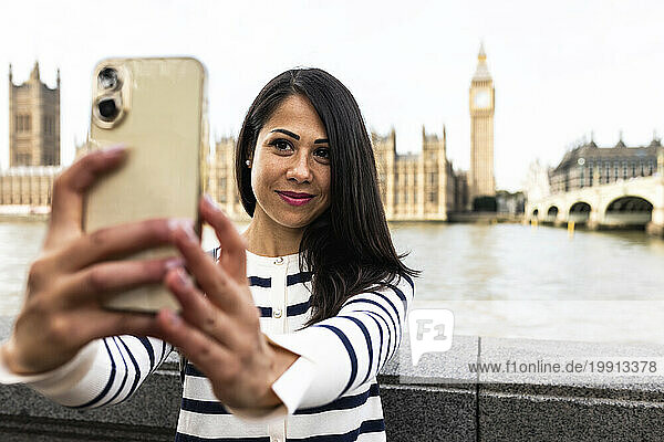Smiling woman taking selfie with Big Ben and Houses of Parliament through smart phone in London city