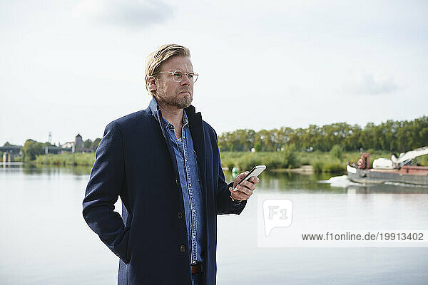 Thoughtful businessman standing with smart phone near river